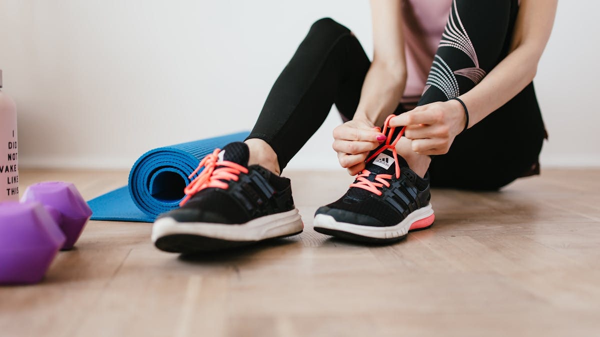 Crop sportswoman in sportswear tying sneakers while sitting on wooden floor with sport mat and dumbbells before exercising indoors